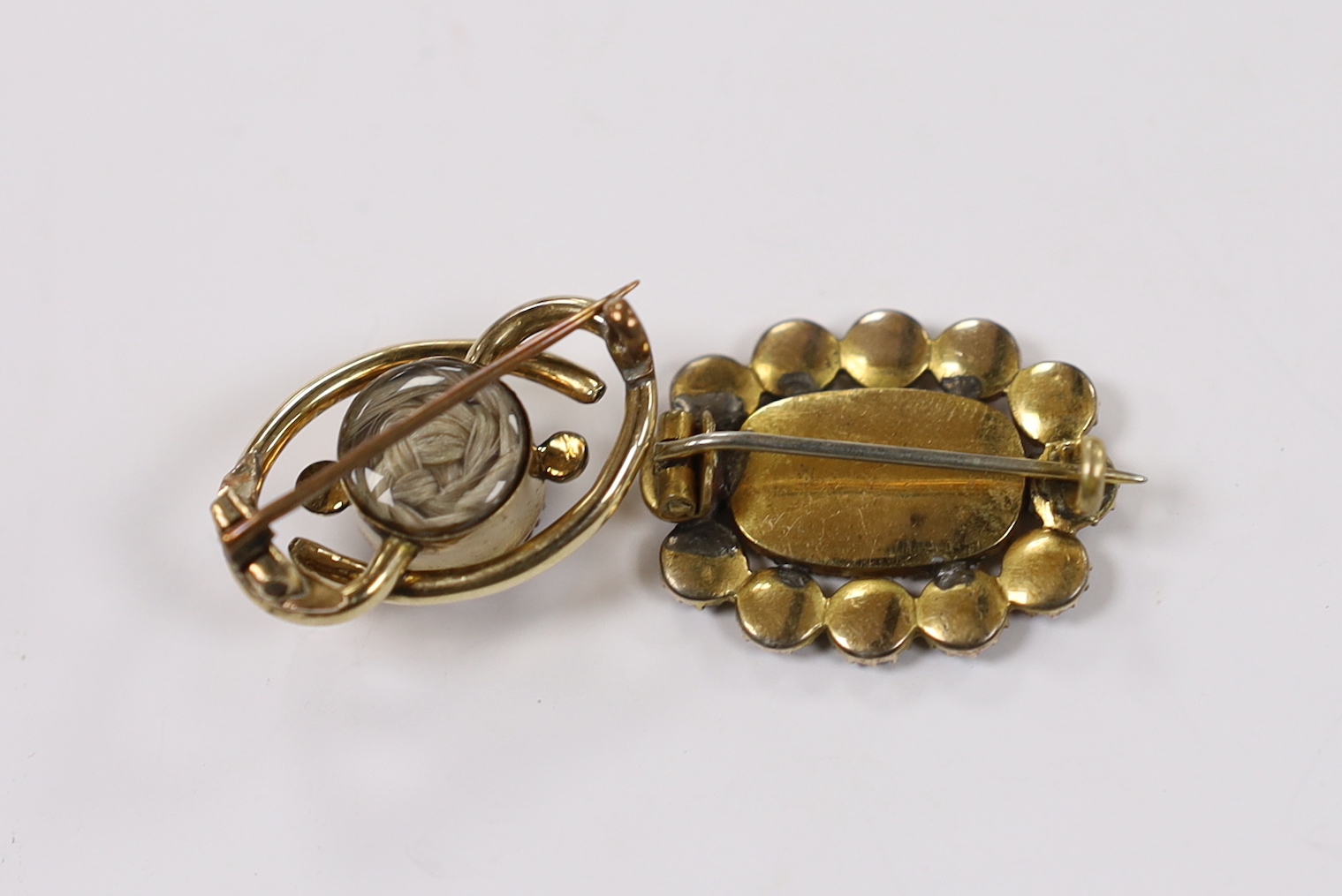 A Victorian yellow metal and single stone foil backed sapphire set scrolling mourning brooch, verso with hair beneath a glazed panel, 26mm, together with a Regency yellow metal, garnet and seed pearl cluster set mourning